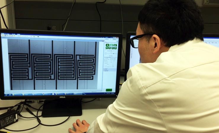 <p>A researcher in the laboratory of Fatih Sarioglu watches ovarian cancer cells move through a hybrid microfluidic chip. The microfluidic channels are vertical; the circuit pattern is shown behind them. (Credit: John Toon, Georgia Tech)</p>