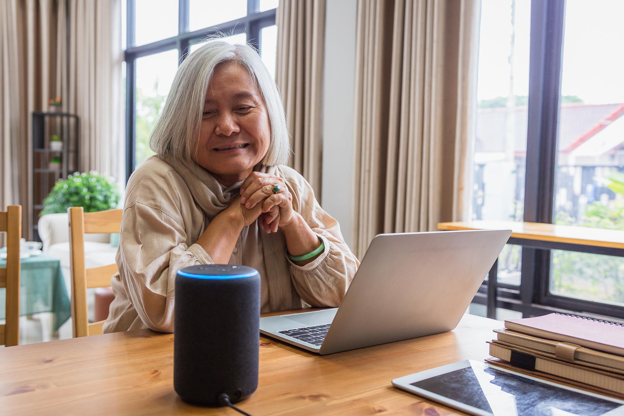 An elderly woman with short white hair smiles and looks at a smart speaker system.