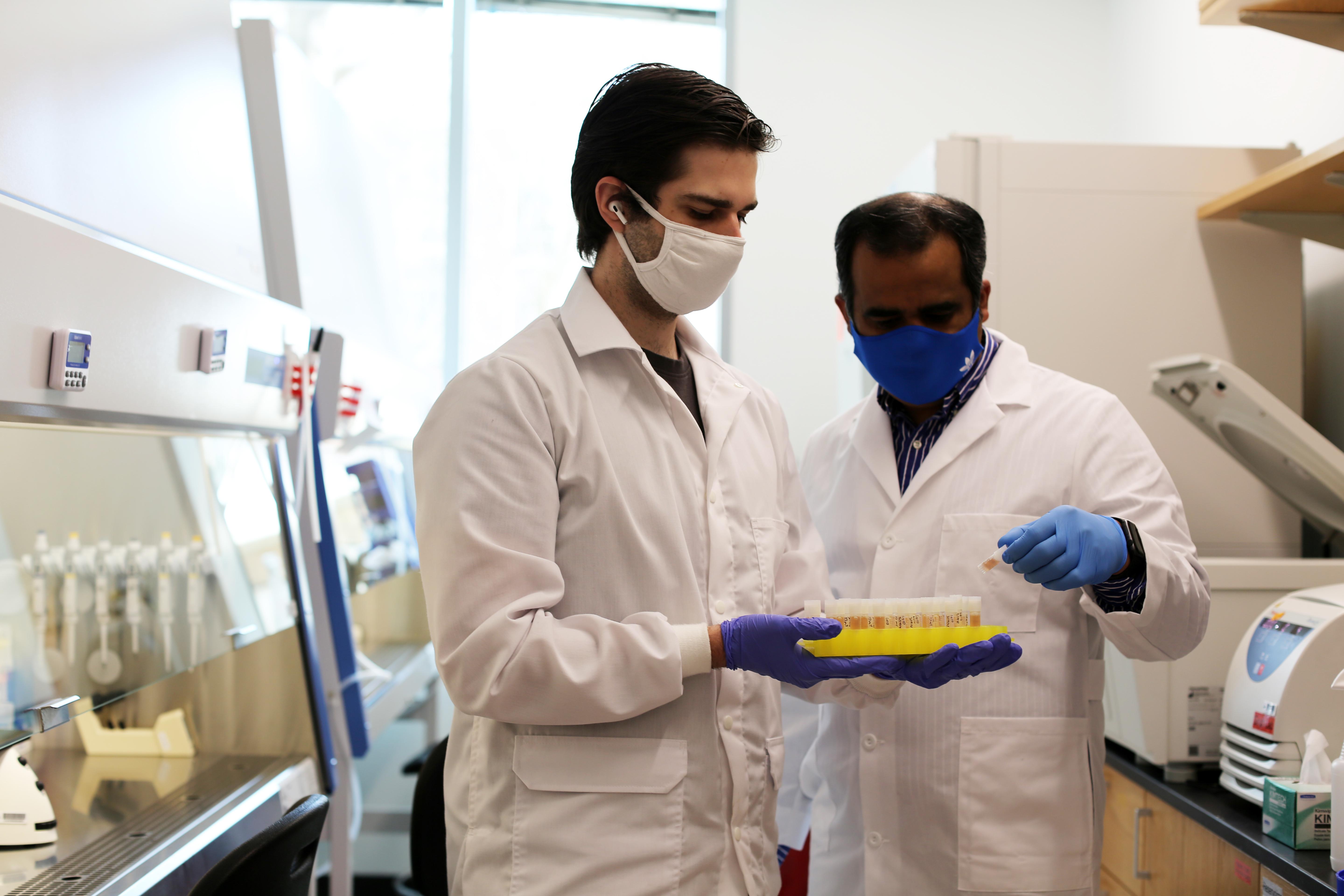 Two people in white lab coats and blue gloves hold a tray of samples. 