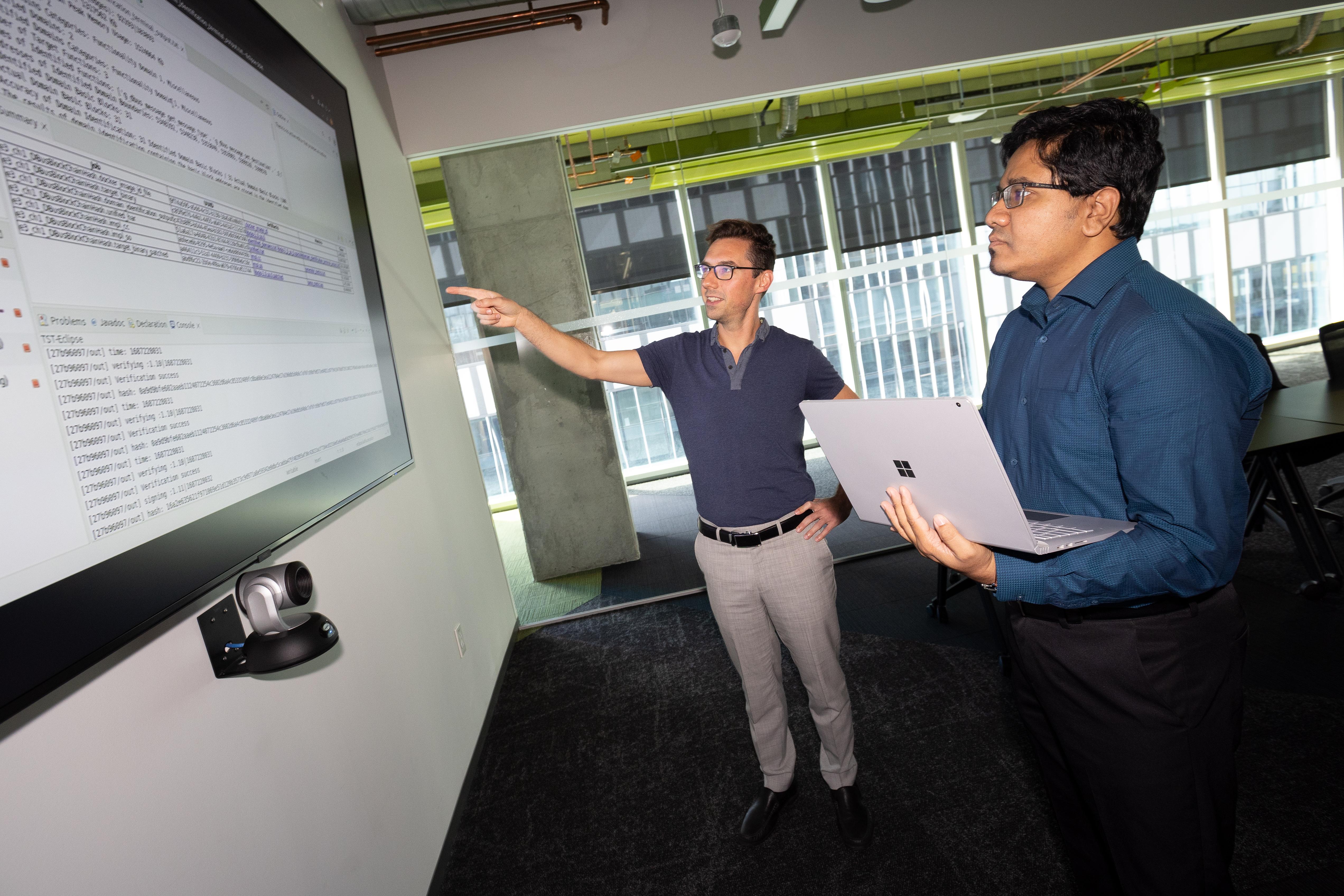 Brendan Saltaformaggio and Amit Sikder stand and look at a large screen displaying computer code. (Photo: Candler Hobbs)